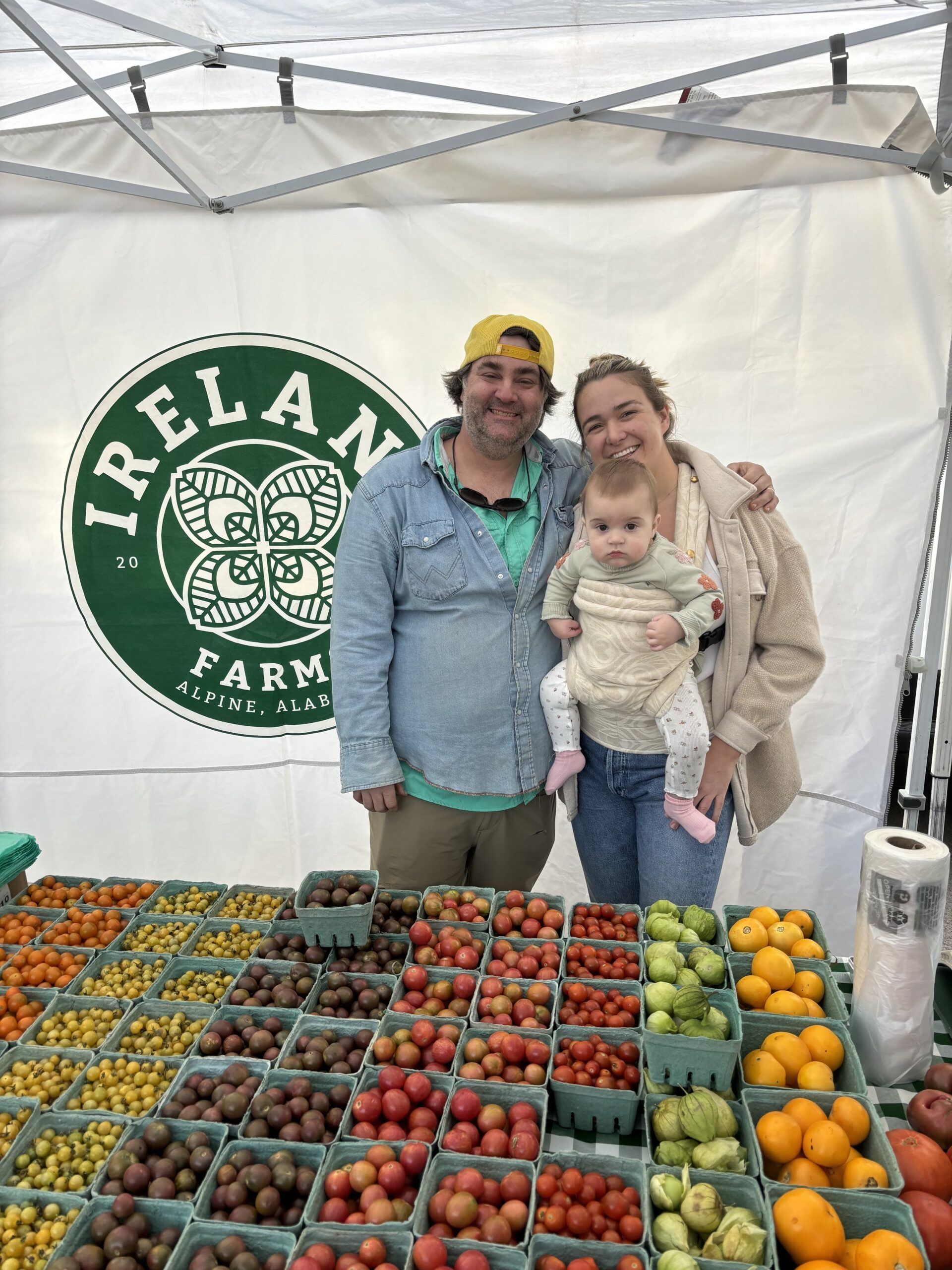Family at a farmers market stand in Alpine, Alabama, showcasing a variety of colorful fresh produce, including tomatoes and citrus fruits, with an "Ireland Farms" banner in the background.